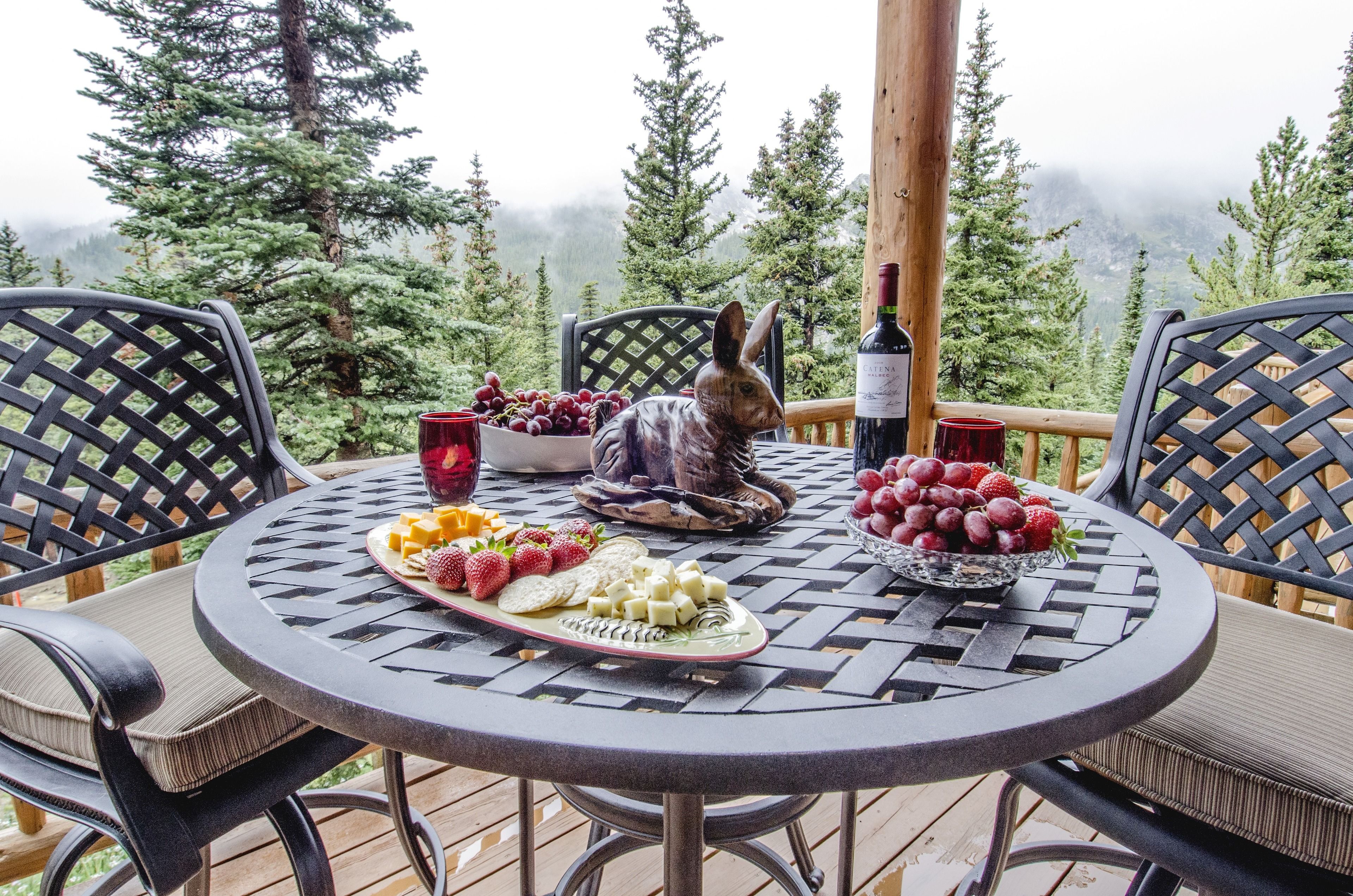 Une table ronde, sur laquelle sont disposés divers fruits et fromages, est entourée de chaises sur une terrasse offrant une vue sur les arbres et les montagnes.