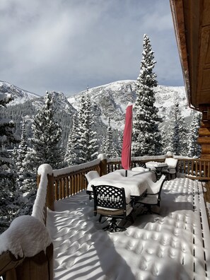 Une terrasse en bois, avec des meubles recouverts de neige et une planche de snowboard rouge appuyée contre la rambarde, offrant une vue sur un paysage montagneux enneigé.