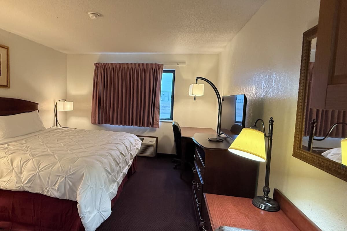 Quarto de hotel com cama, escrivaninha, cadeira, duas luminárias, espelho e janela com cortinas.