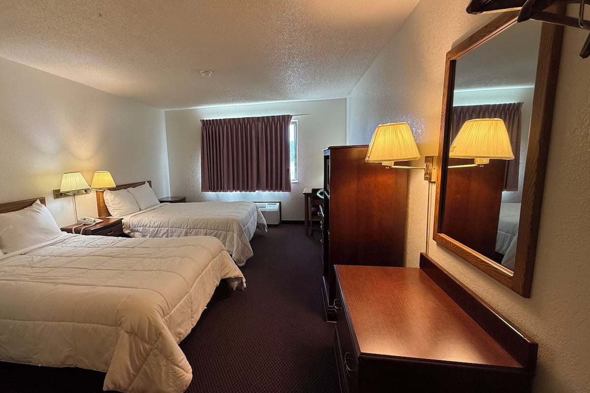 Quarto de hotel com duas camas, uma escrivaninha, uma cadeira, um espelho e uma janela com cortinas.