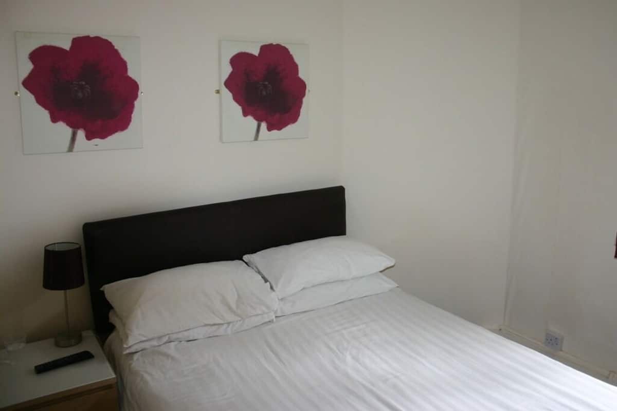 Quarto com cama, roupa de cama branca, cabeceira preta, mesa de cabeceira com abajur e duas peças de arte floral vermelha na parede.