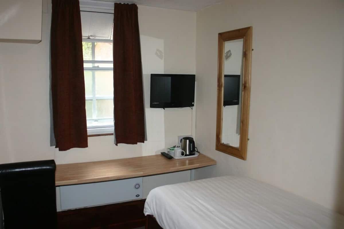 Quarto de hotel com cama, escrivaninha com cadeira, televisão na parede e janela com cortinas.