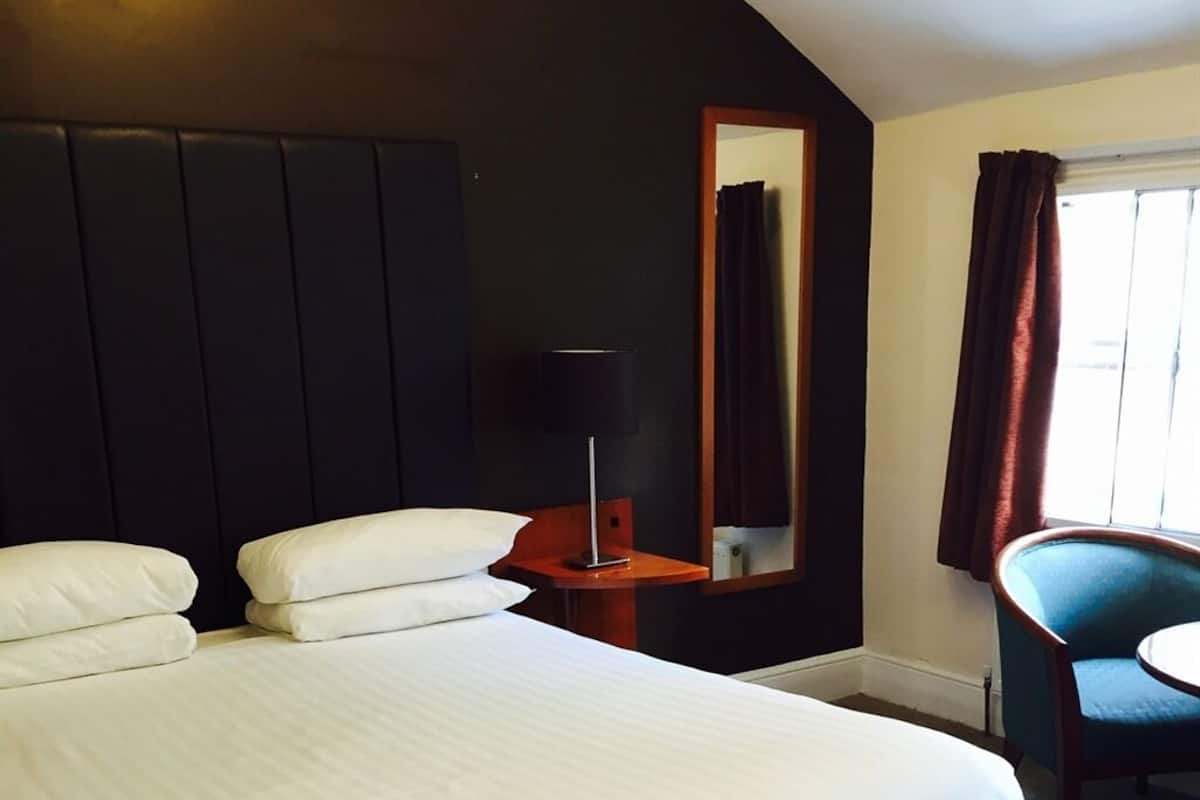 Quarto de hotel com uma cama grande, uma mesa com luminária, uma cadeira e uma janela com cortinas.