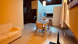 A modern living room with a sofa, dining table, and chairs, a kitchen area in the background, and a large window.
