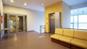 Un área de espera moderna con un sofá amarillo, un ascensor y una entrada por puerta de cristal.