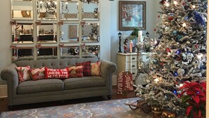 A cozy living room with a grey sofa, a chandelier, a Christmas tree, and a patterned rug.