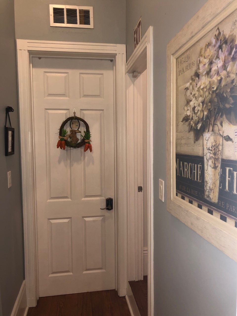 A hallway with a white door, a wall-mounted vent, and a framed artwork depicting flowers and text.