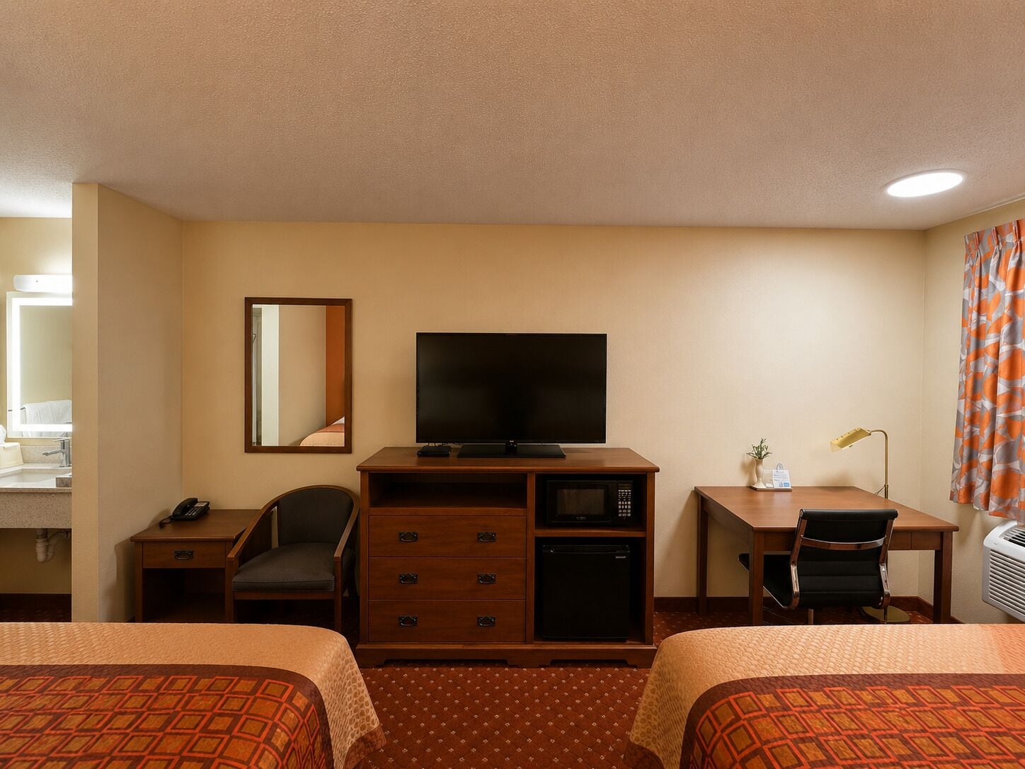 A hotel room with two beds, a desk with a television, a chair, a desk lamp, and a mirror.