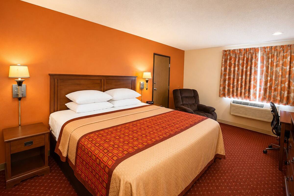 A hotel room with a large bed, bedside lamps, a chair, an air conditioning unit, and a door.
