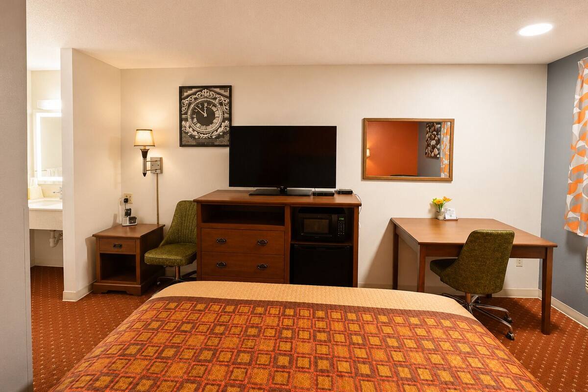 A hotel room with a bed, a desk, a television, a chair, a lamp, and a clock on the wall.