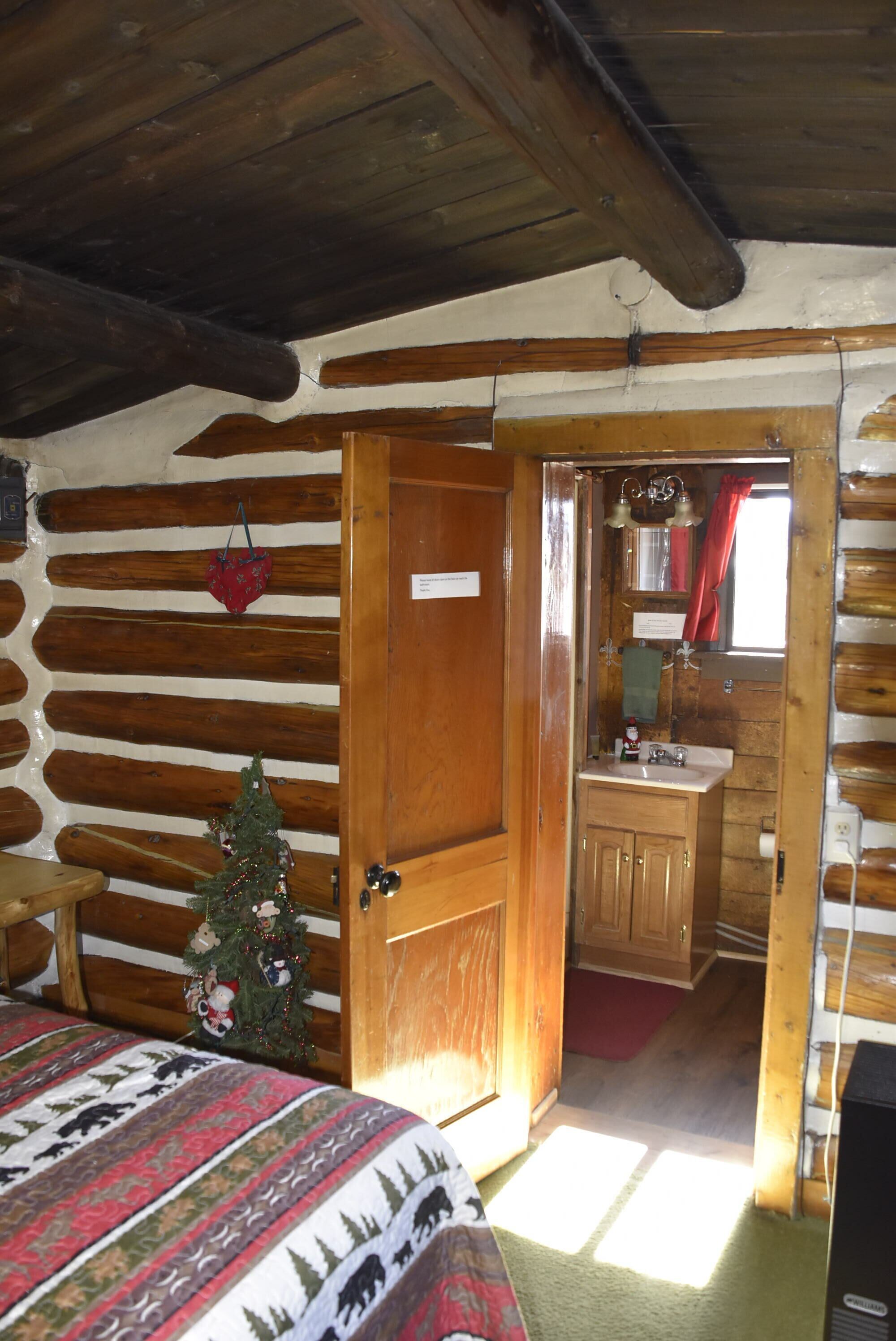 A log cabin bedroom with a wooden door, a small Christmas tree, a bed with a patterned blanket, and a wooden bench.