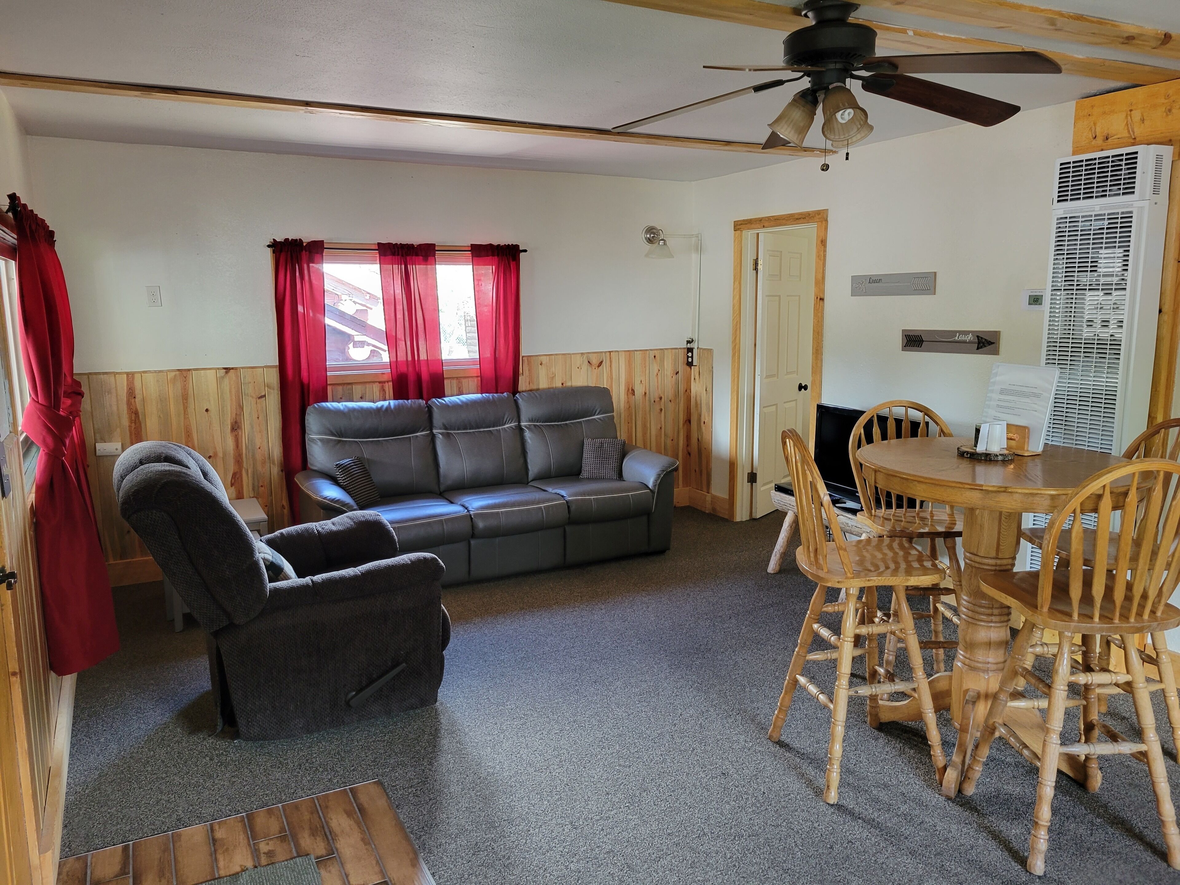 A cozy room with a leather sofa, a wooden dining table with chairs, a ceiling fan, and a window with red curtains.