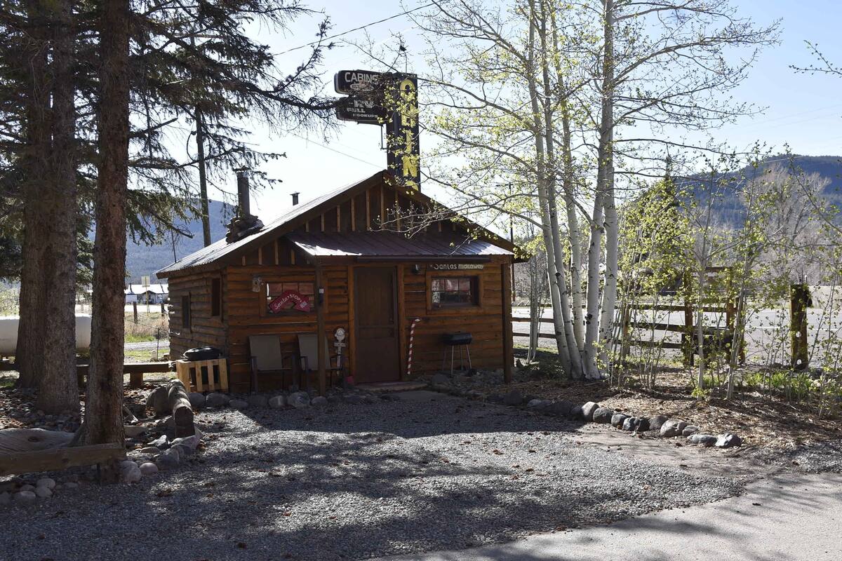 Une cabane en bois rustique, dotée d’une cheminée, entourée d’arbres et bordée d’un chemin de gravier.