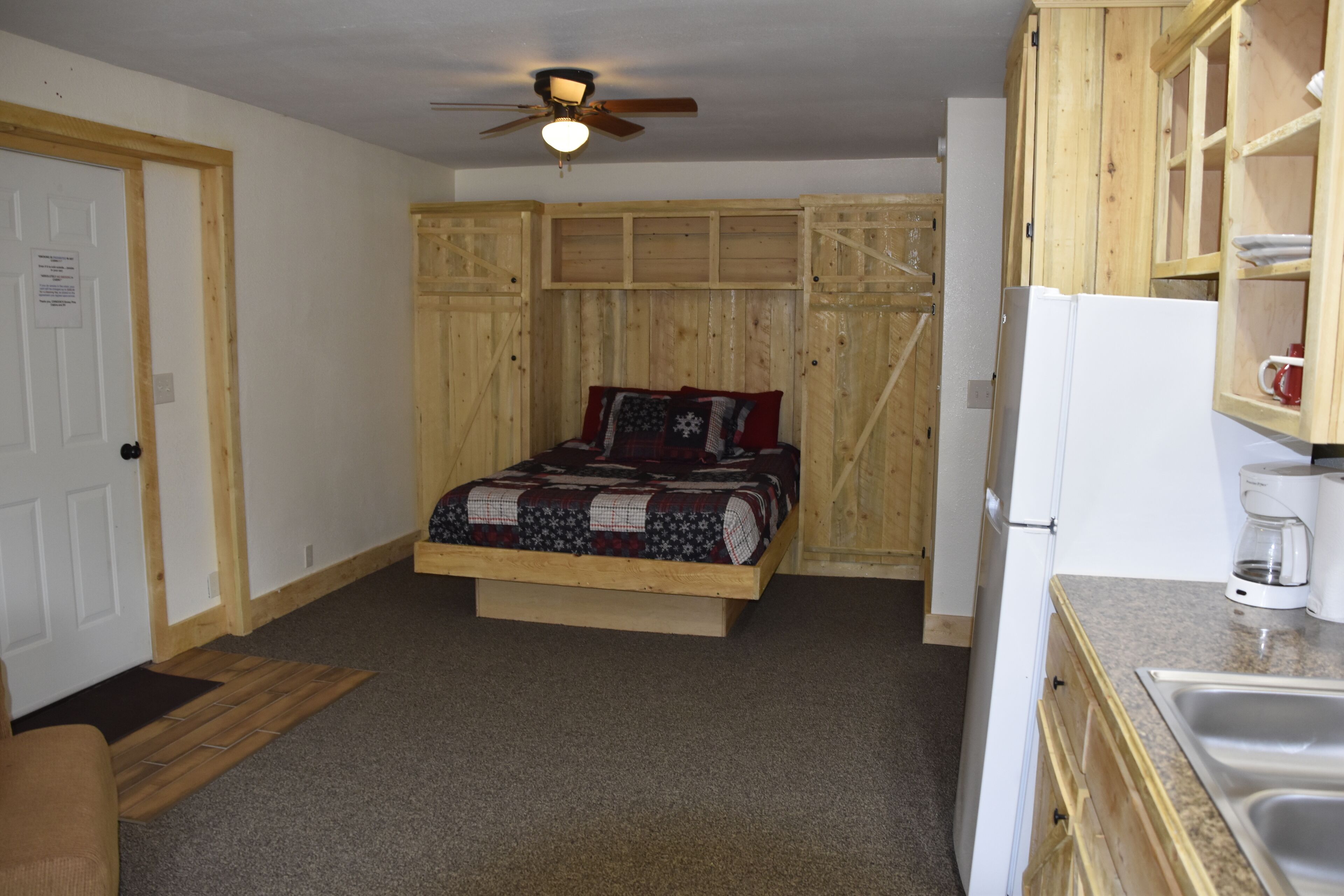 Une chambre avec un lit en bois, un ventilateur de plafond et une kitchenette équipée d’un réfrigérateur et d’une machine à café.