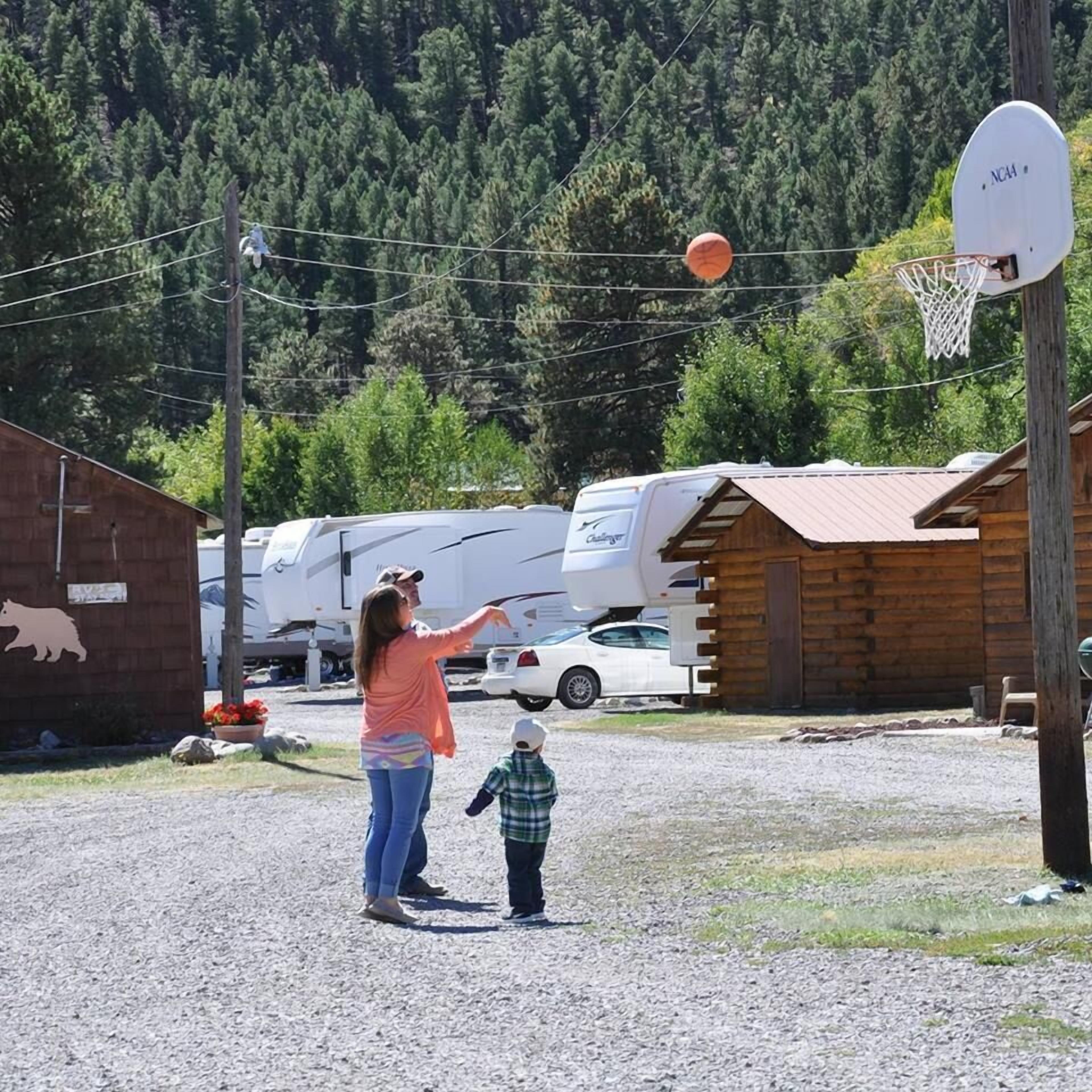 A woman and a child playing basketball in a gravel area with RVs and a wooden cabin in the background.