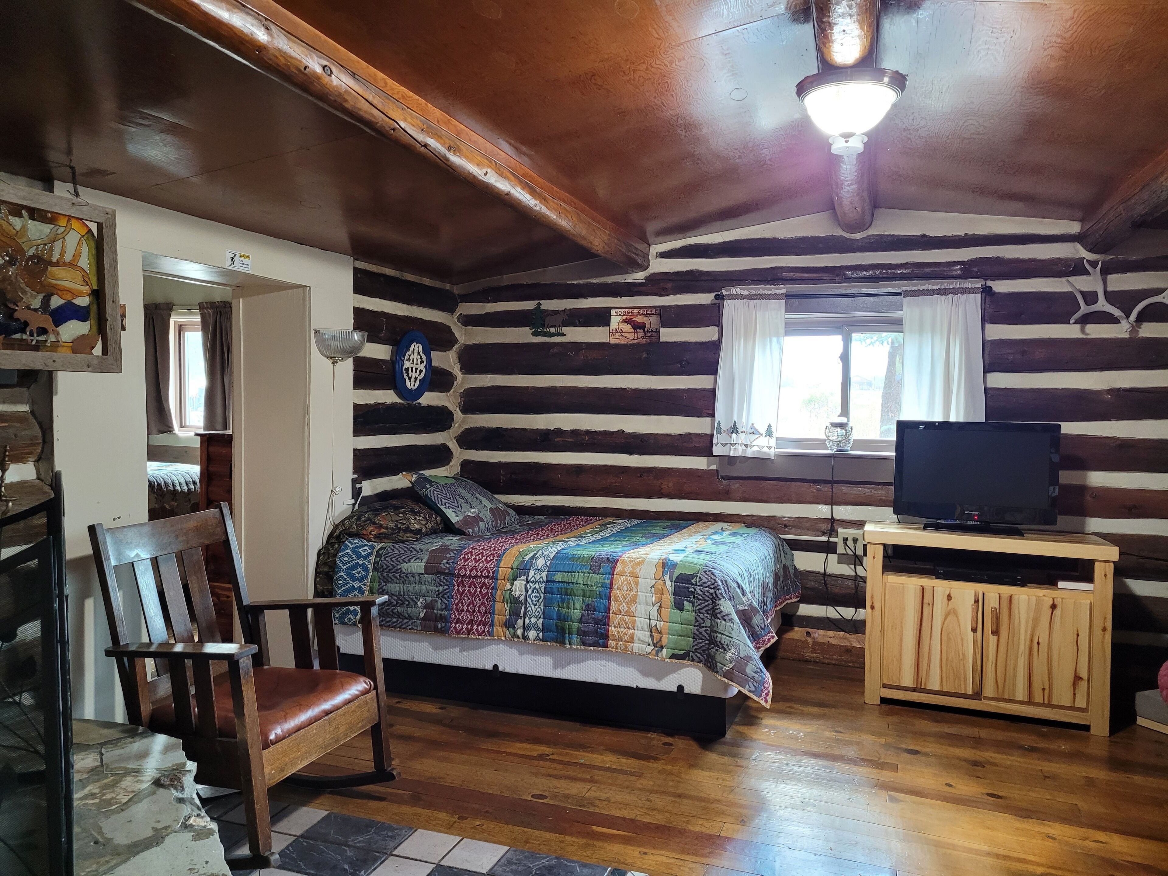 Una habitación tipo cabaña de madera con una cama, una silla de madera, un televisor sobre un mueble de madera y una ventana con cortinas.