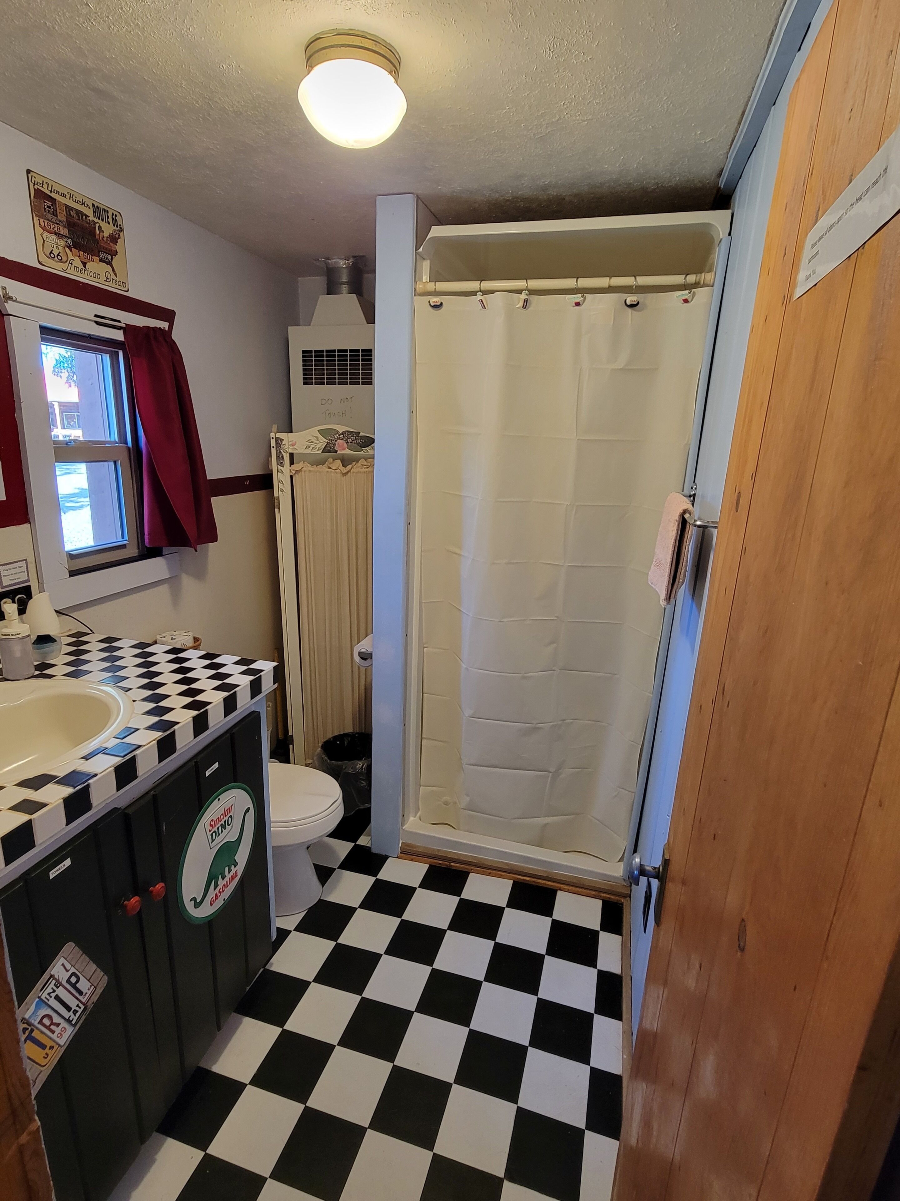 A bathroom with a black and white checkered floor, a toilet, a shower with a curtain, a sink, and a window with a red curtain.
