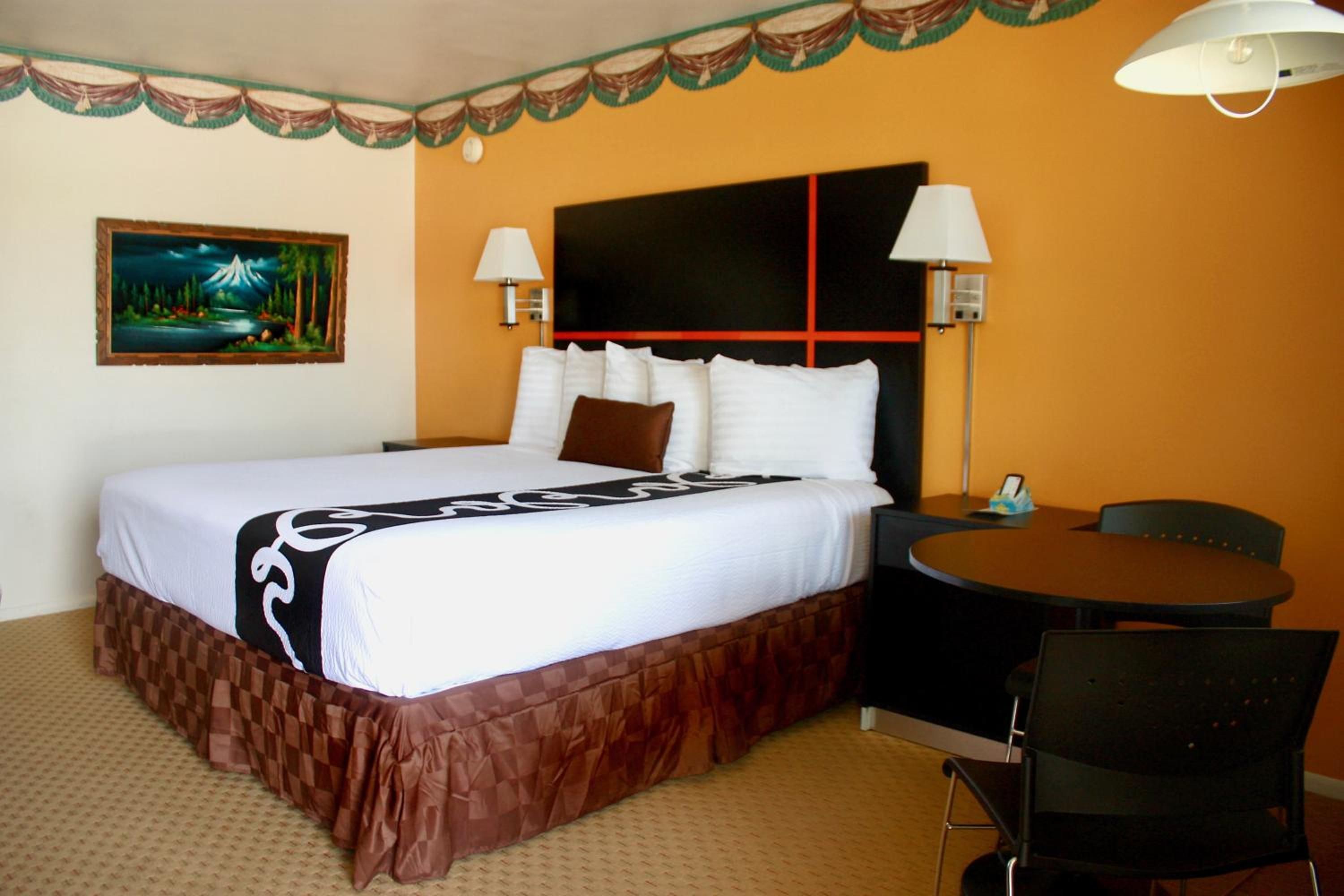 Habitación de hotel con una cama grande, dos lámparas de noche, una mesita redonda con una silla y un cuadro en la pared.