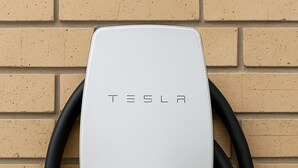 A white Tesla charging station with a green light, connected to a brick wall. - Chaparral Motor Inn (Burlington)