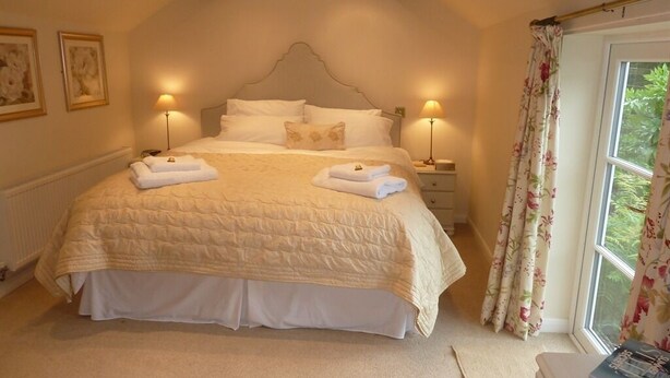 A neatly made bed with a quilted cover, two bedside lamps, and a window with floral curtains. - The Coach House (Stafford)