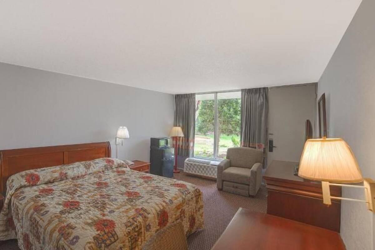 A hotel room with a bed, a chair, a desk, a lamp, and a window with curtains.