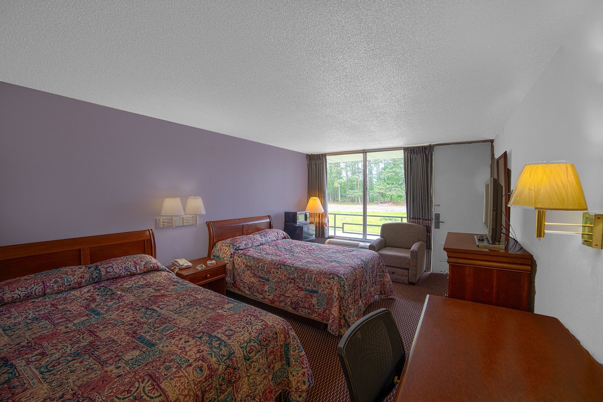 A hotel room with two beds, a desk, a chair, a TV, and a window with a view of greenery.