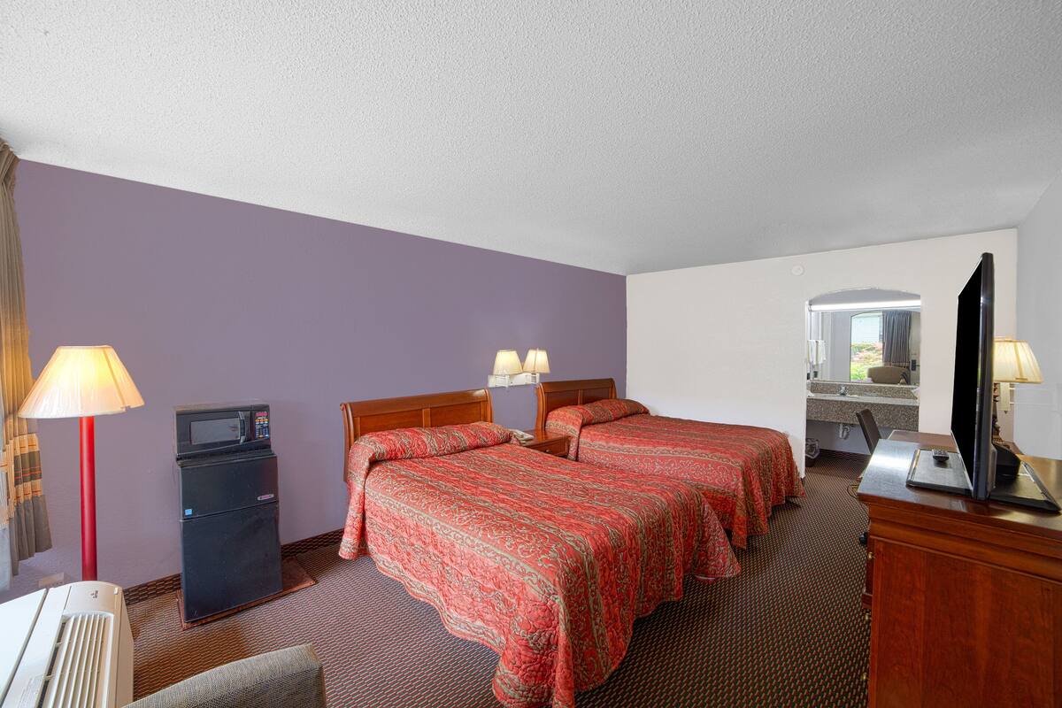 A hotel room with two beds, a refrigerator, a desk, a television, and a bathroom with a sink and mirror.