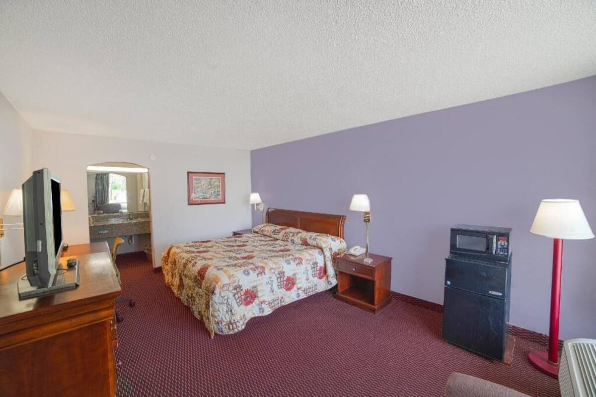 A hotel room with a bed, a desk with a television, a desk lamp, a nightstand, a refrigerator, and a wall-mounted mirror.