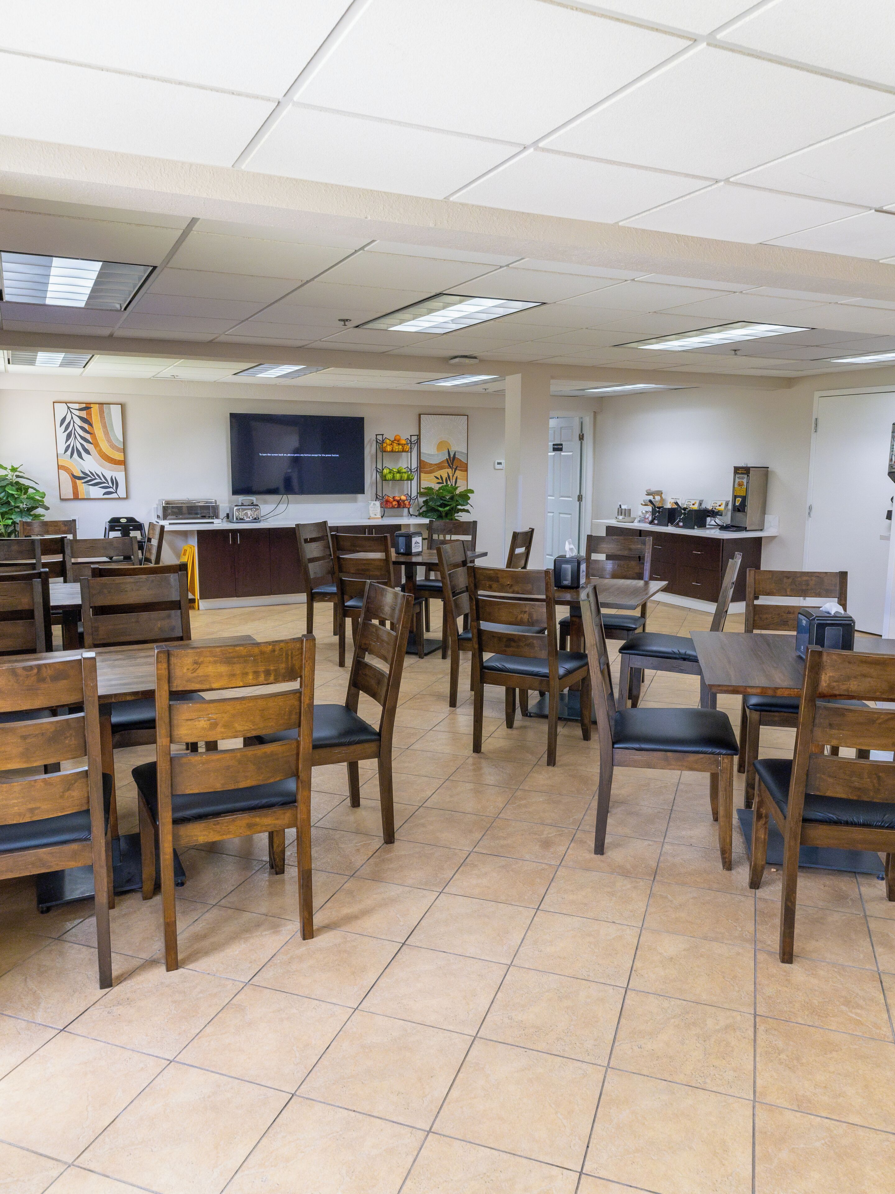 Un espace repas avec plusieurs tables et chaises, un coin cuisine équipé d’appareils électroménagers et un téléviseur à écran plat fixé au mur.