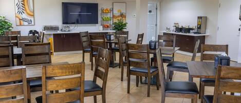 Un espace repas avec plusieurs tables et chaises, un coin cuisine équipé d’appareils électroménagers et un téléviseur à écran plat fixé au mur.