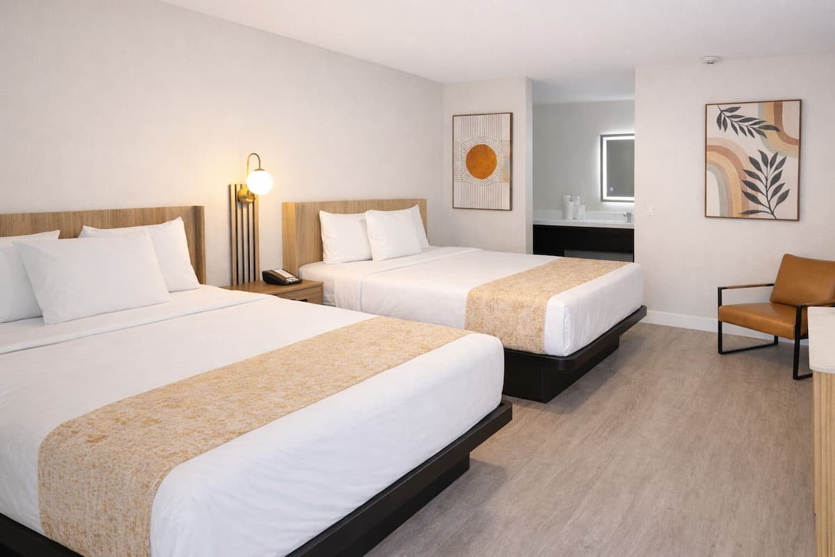 A hotel room with two beds, a desk, a chair, a nightstand with a lamp, and artwork on the walls.
