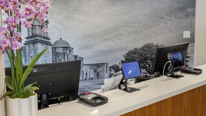 A reception desk with two computer monitors, a potted plant, and a large wall mural. - The Alcala Hotel Mission Valley/Seaworld (San Diego)