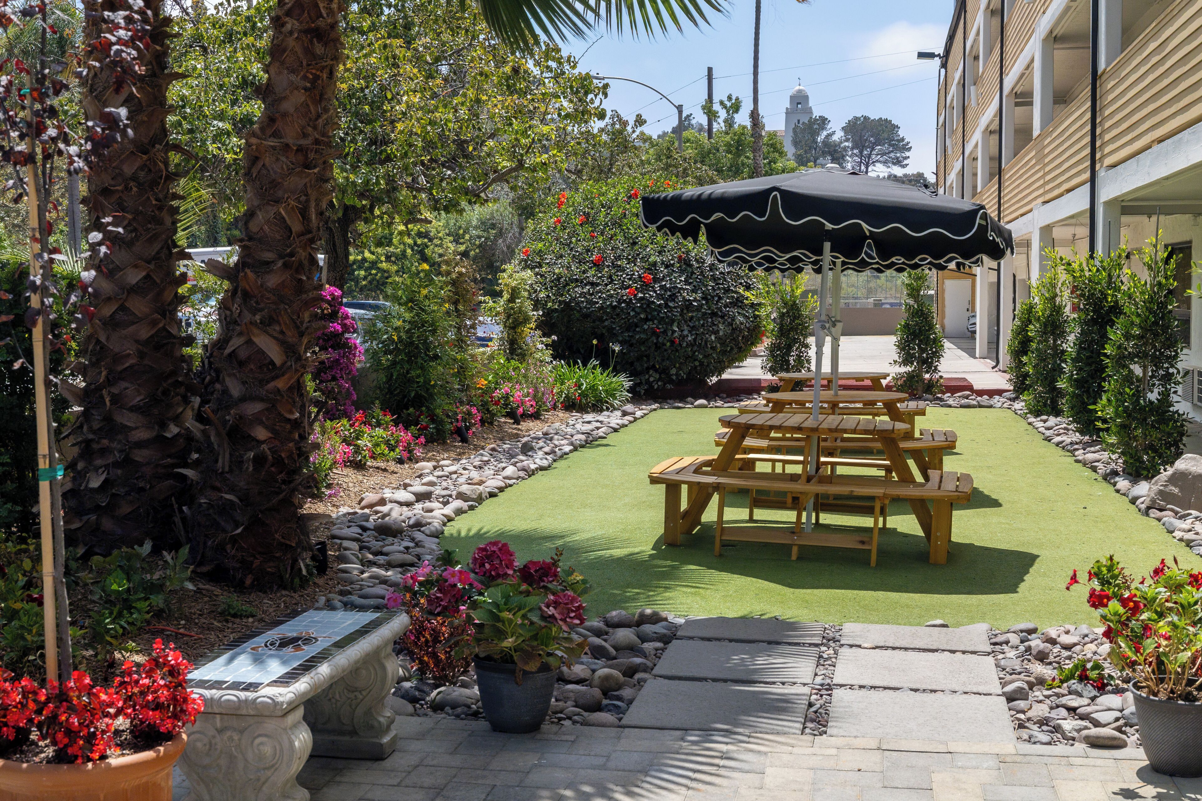 A patio area with a wooden picnic table, a black umbrella, and a variety of potted plants and flowers.