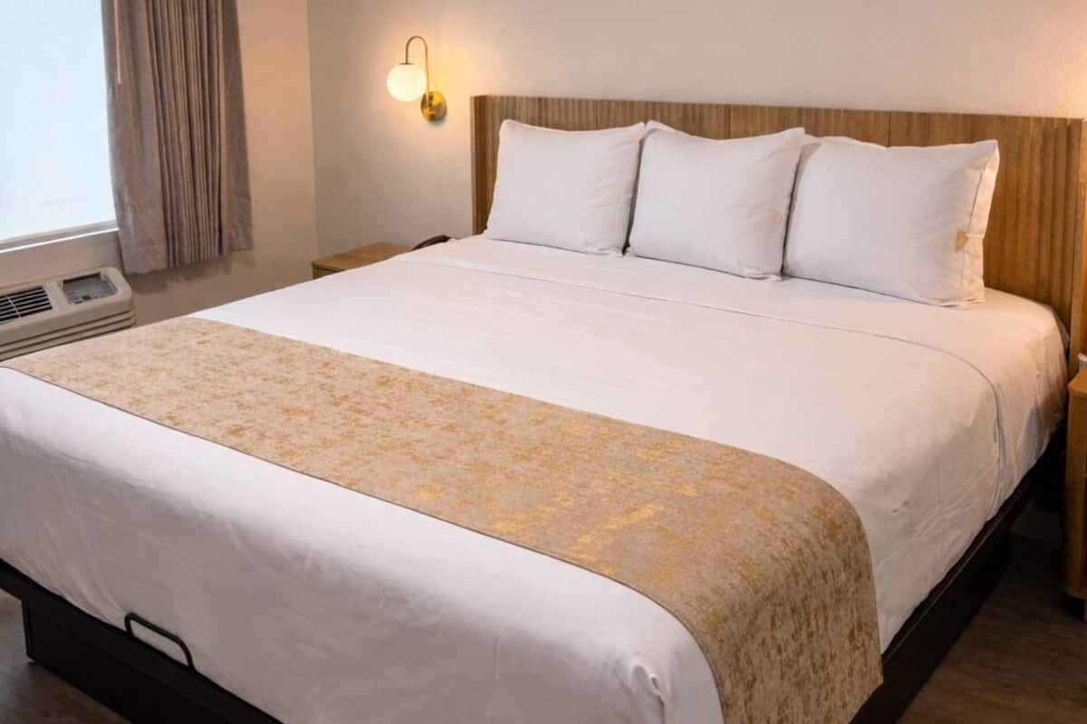 A hotel room with a large bed, a bedside table, a wall-mounted lamp, and a window with curtains.
