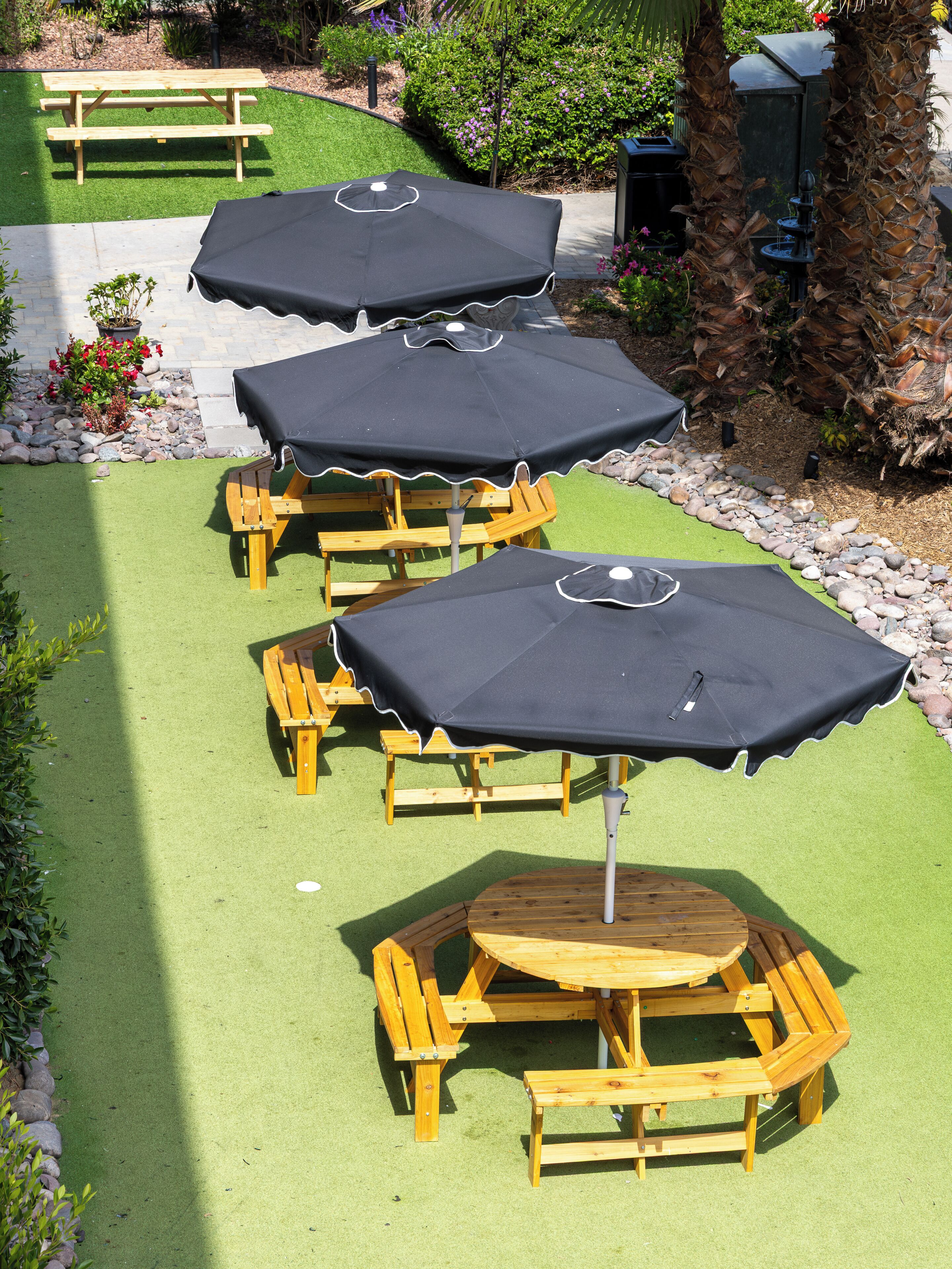 A patio area with multiple wooden tables and black umbrellas, surrounded by greenery and a stone pathway.
