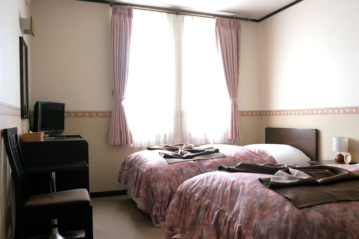 Une chambre d’hôtel avec deux lits, un bureau avec un ordinateur, une chaise et une table de chevet avec une lampe et une horloge.