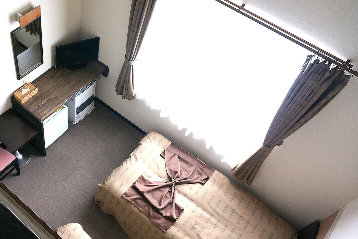 Une chambre à coucher comprenant un lit, un bureau, une chaise, un téléviseur et une fenêtre avec des rideaux.