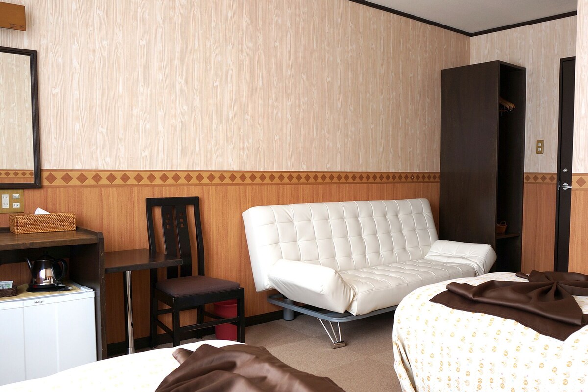 Une chambre d’hôtel avec un lit, un fauteuil blanc, un bureau avec une bouilloire et un petit réfrigérateur.