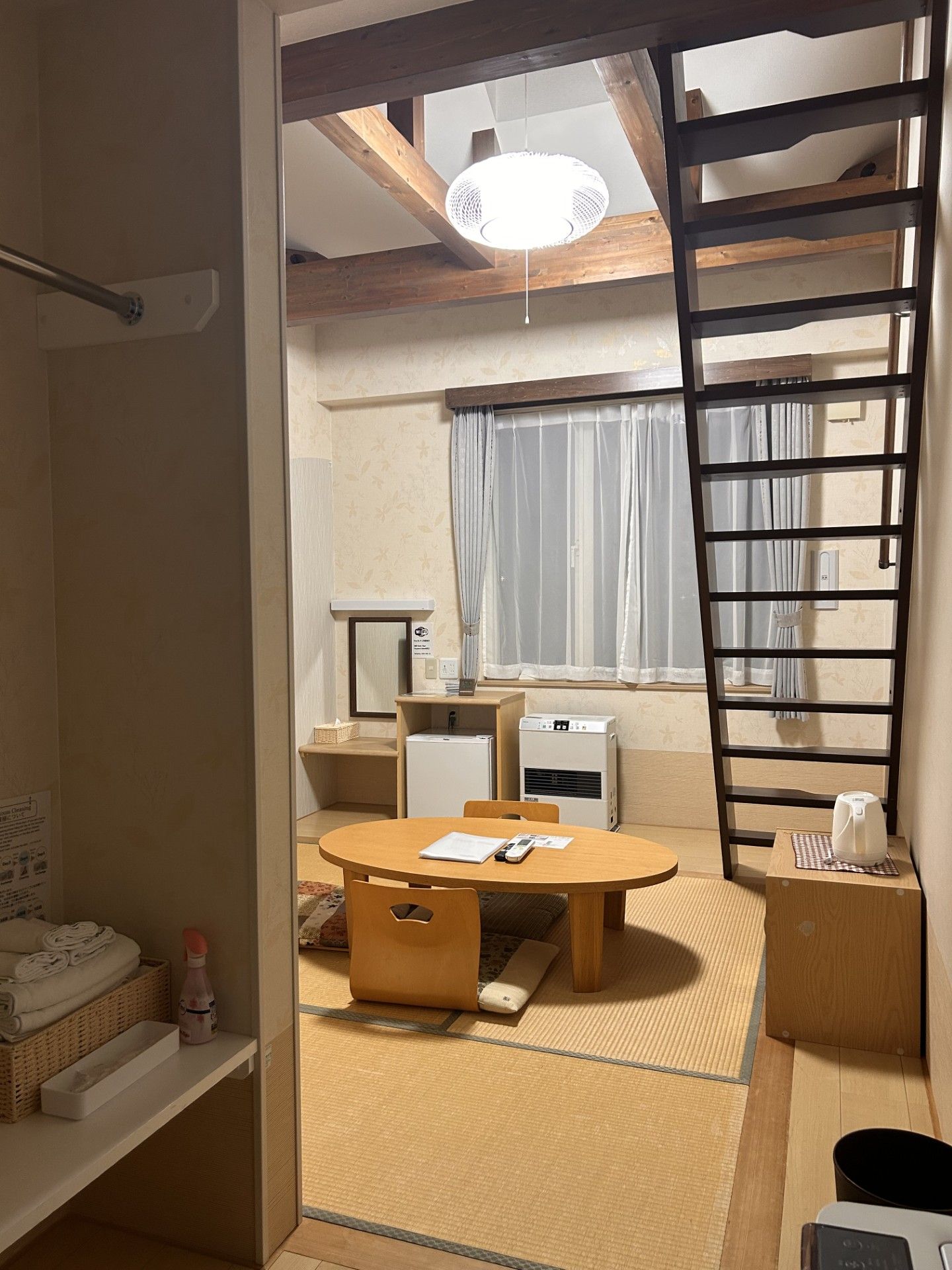 Habitación de hotel compacta con techo de madera, una mesa redonda para comer, una pequeña cocina y un ventanal con cortinas.
