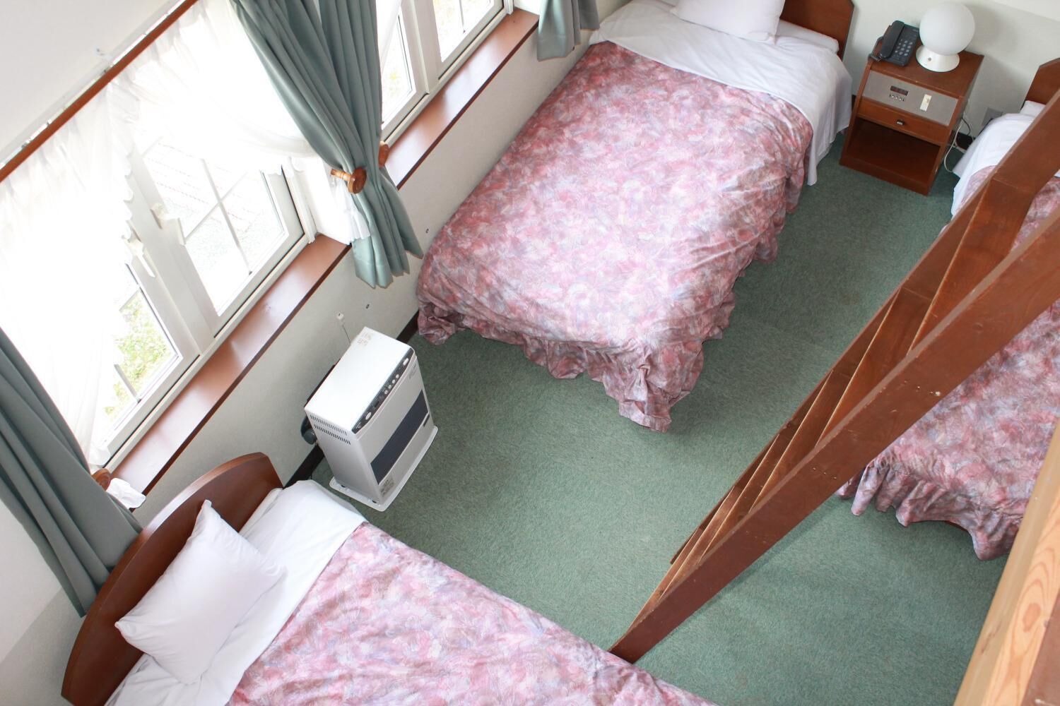 A hotel room with two beds, a bedside table with a lamp, a heater, and a window with curtains.