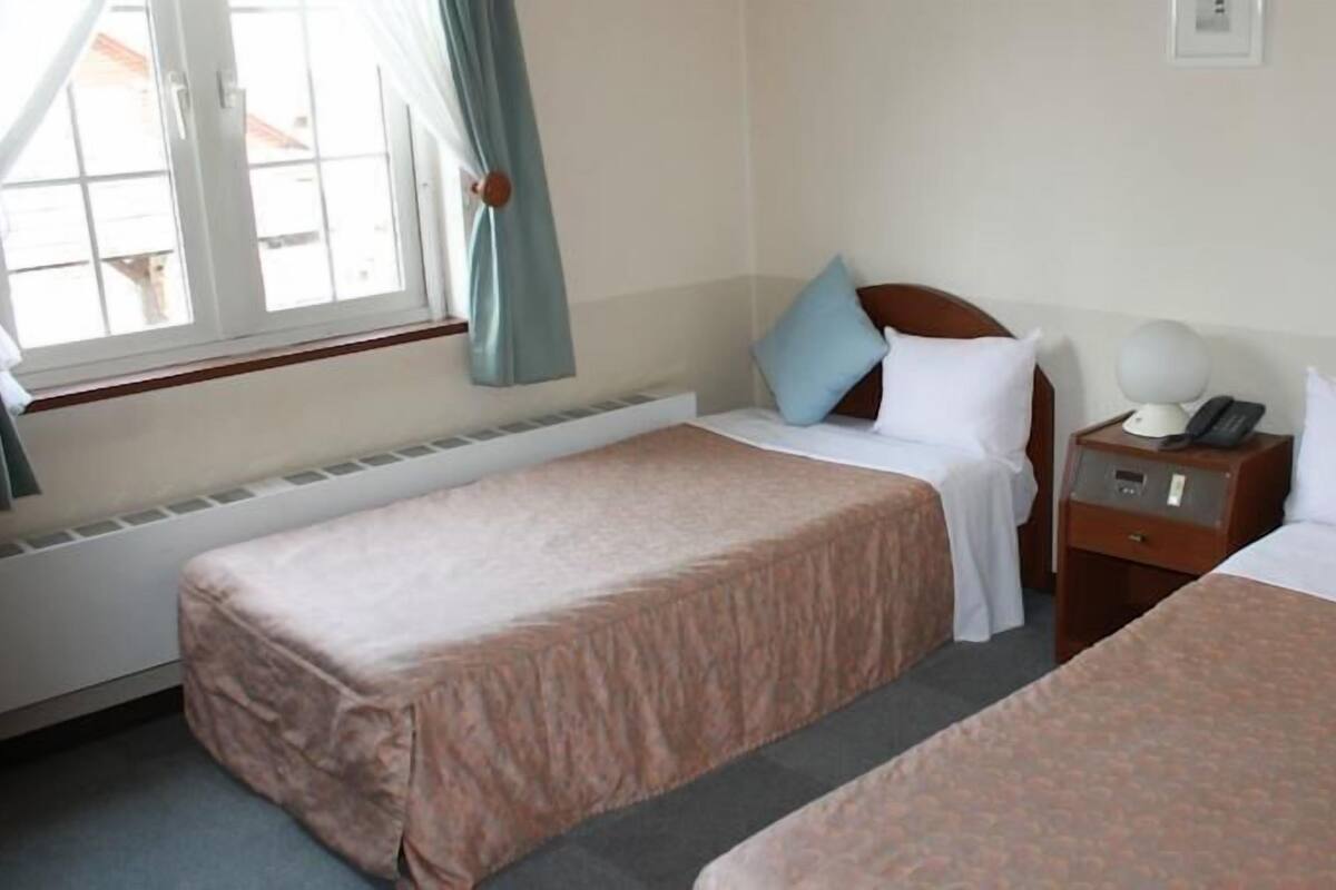 Ein Hotelzimmer mit zwei Einzelbetten, einem Nachttisch mit Lampe und einem Fenster mit Vorhängen.