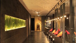 A corridor with a glass wall on one side, a reception desk with a vase of flowers, and several wicker chairs with red cushions. - Atami Tensui (Atami)