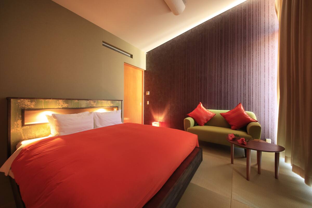 Una habitación de hotel con una cama grande, un sofá, una mesita y una ventana con cortinas.