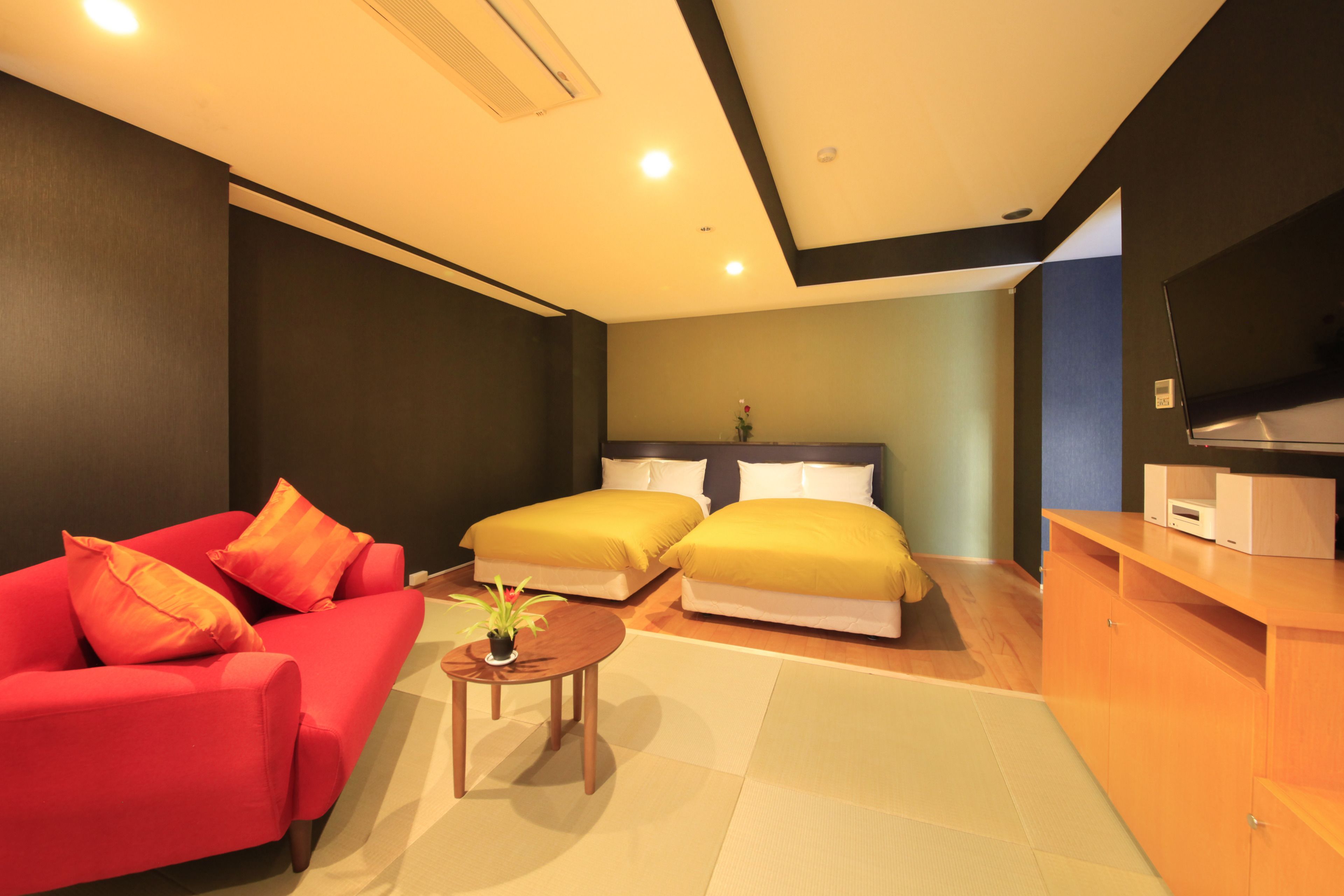 A hotel room with a red sofa, two beds with yellow bedding, a small wooden table with a plant, and a TV on a wooden cabinet.