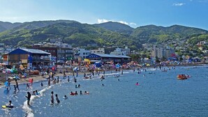 A beach with people swimming and sunbathing, buildings in the background, and mountains in the distance. - Atami Tensui (Atami)