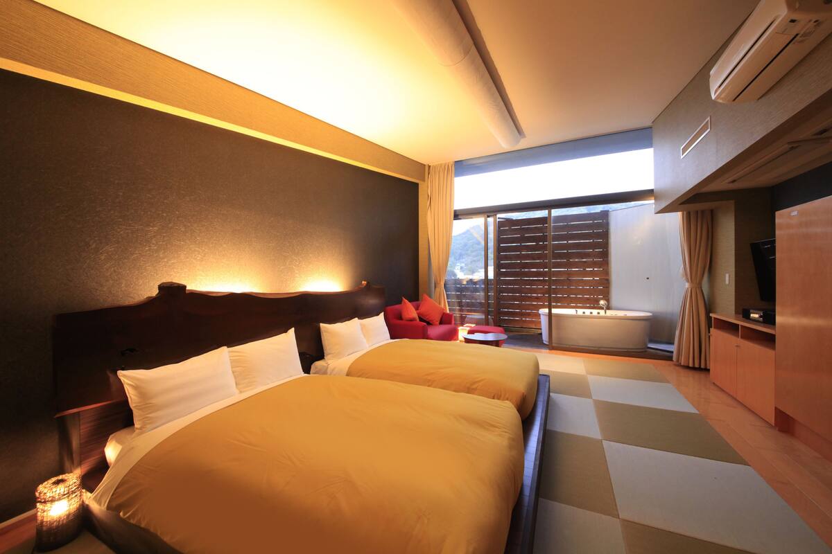 Una habitación de hotel con dos camas, una silla roja, una mesita pequeña y un ventanal grande con persianas.