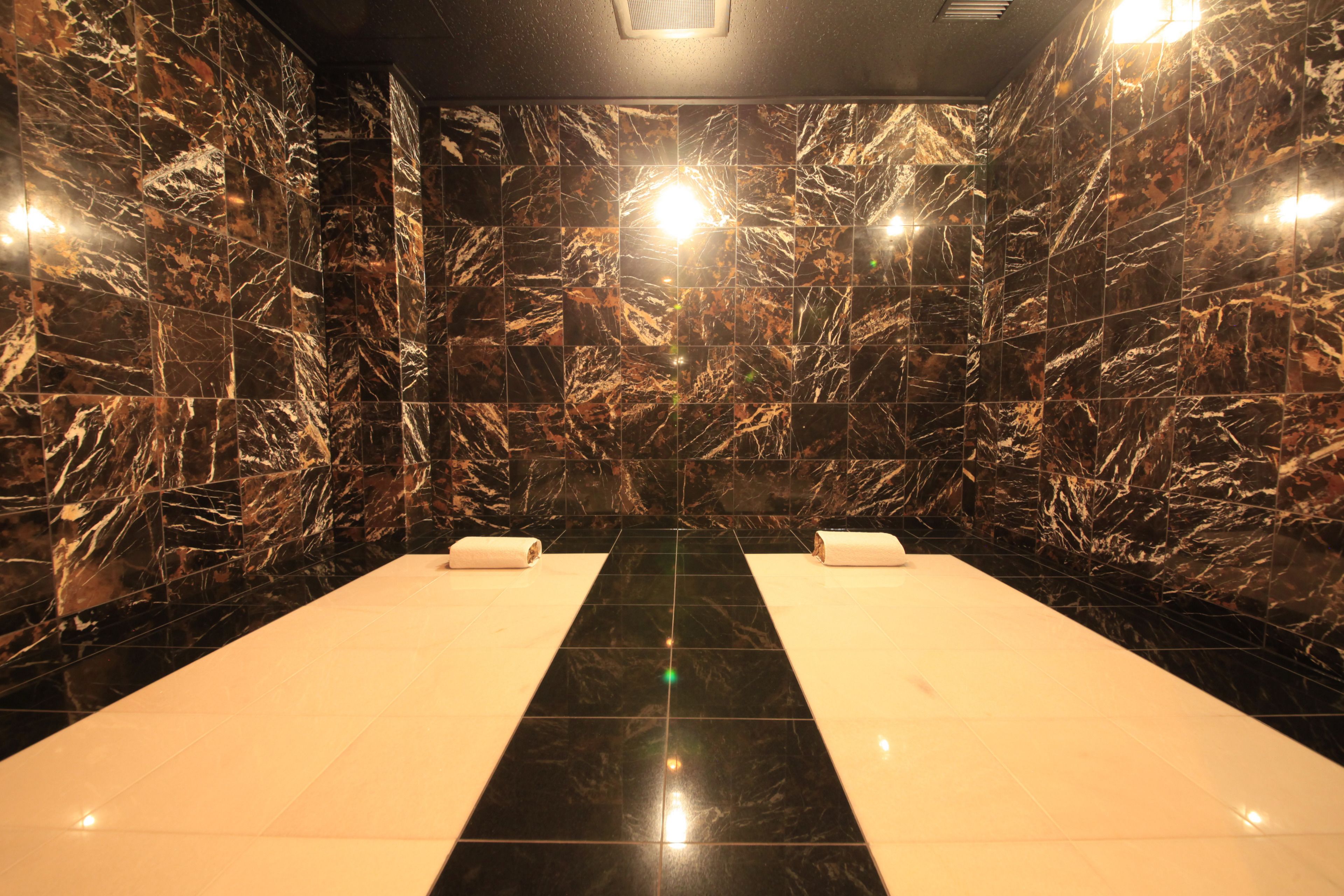 Un cuarto de spa con paredes de mármol negro, un suelo negro reflectante en el centro y dos toallas blancas sobre bancos blancos.