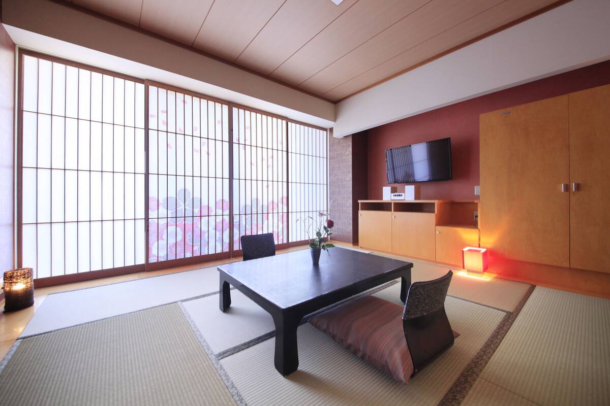 Una habitación tradicional japonesa con suelo de tatami, una mesa baja con cojín, televisor montado en la pared y puertas correderas con vistas a los cerezos en flor.
