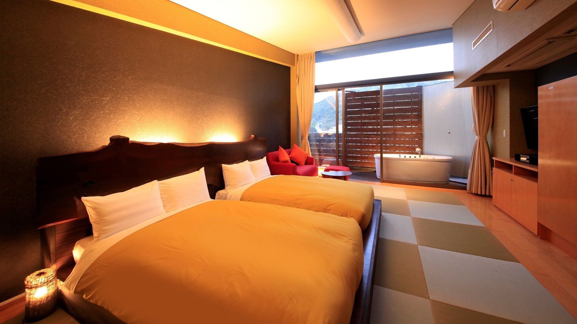 A hotel room with two beds, a sitting area with a red chair, a bathtub, and a large window with a view.