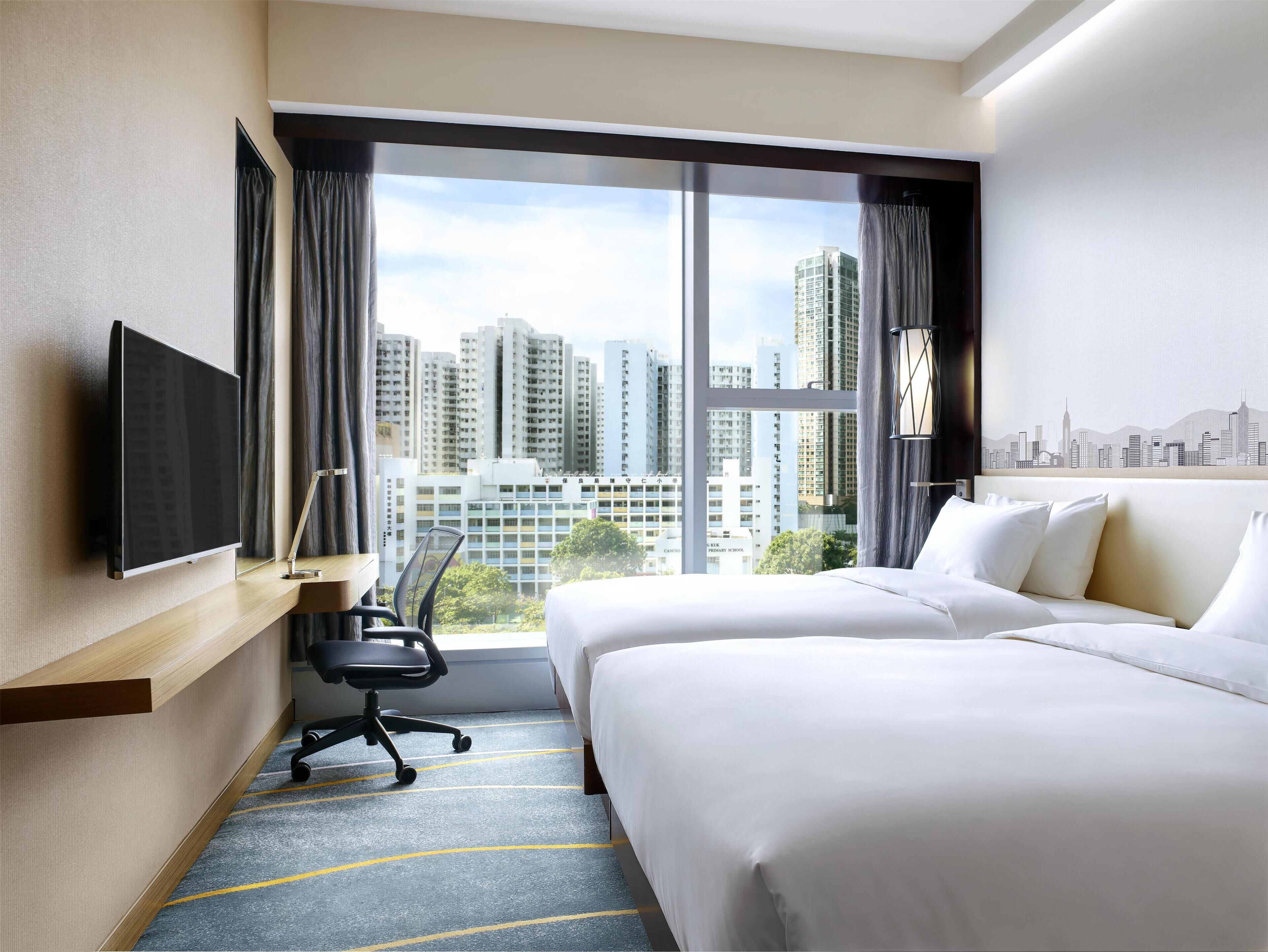 Habitación de hotel con dos camas, un escritorio, una silla, un televisor y un ventanal con vistas a la ciudad.